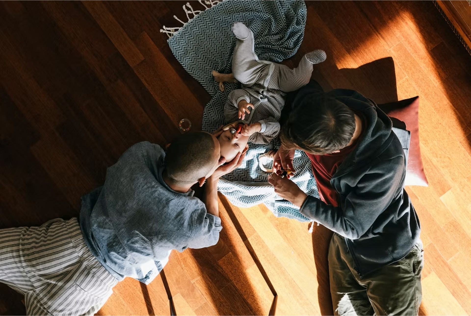 Two parents sit on a warm wooden floor and play with their baby on a patterned blanket